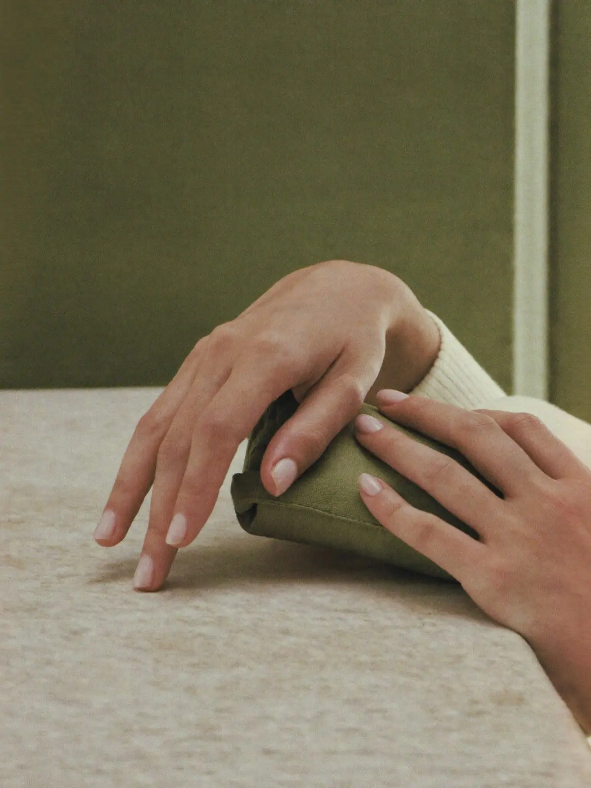 Conceptual campaign image: relaxed hands with sheer nude nails resting on limestone and olive linen