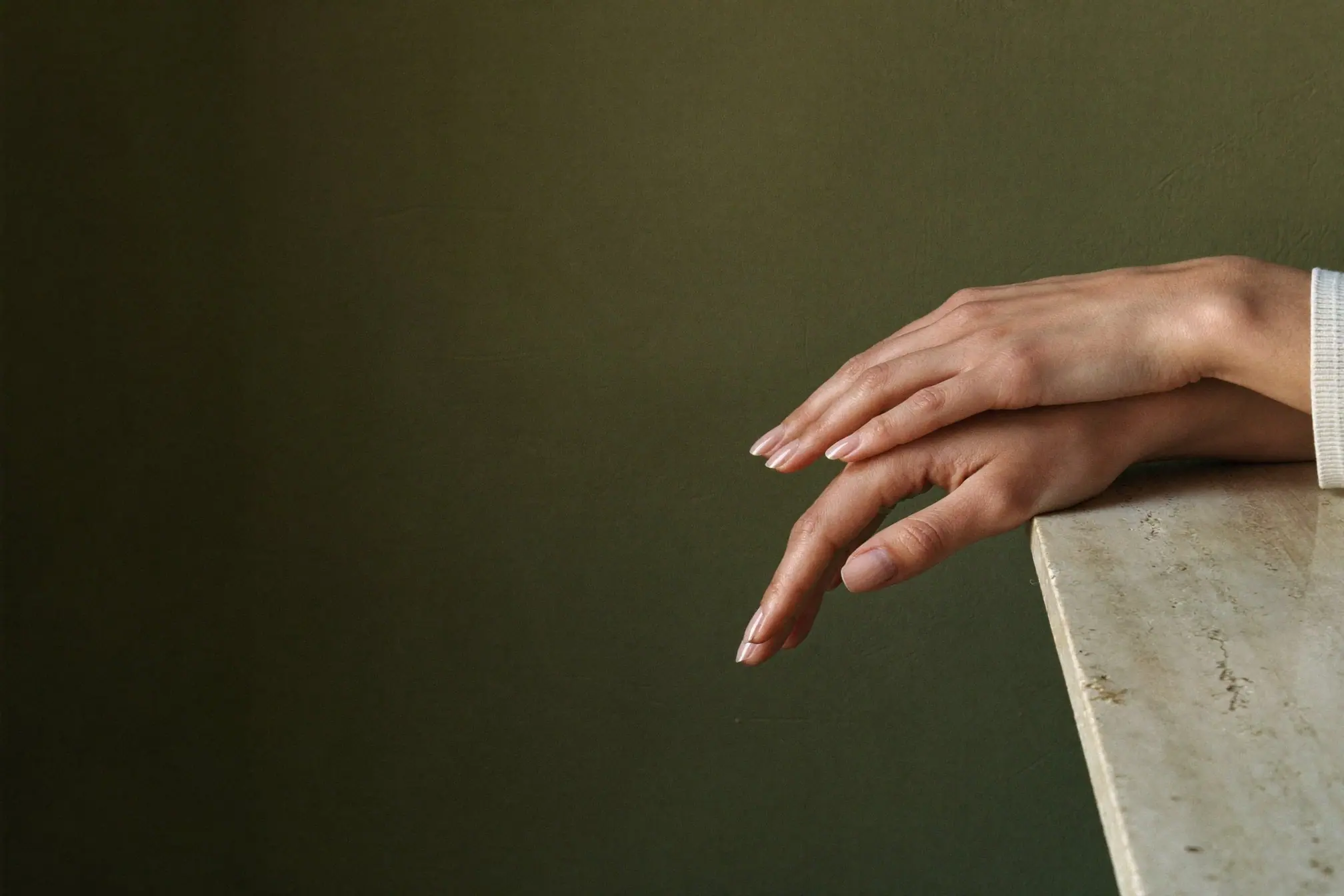 Conceptual campaign image: relaxed hands with sheer nude nails resting on a travertine ledge against an olive wall