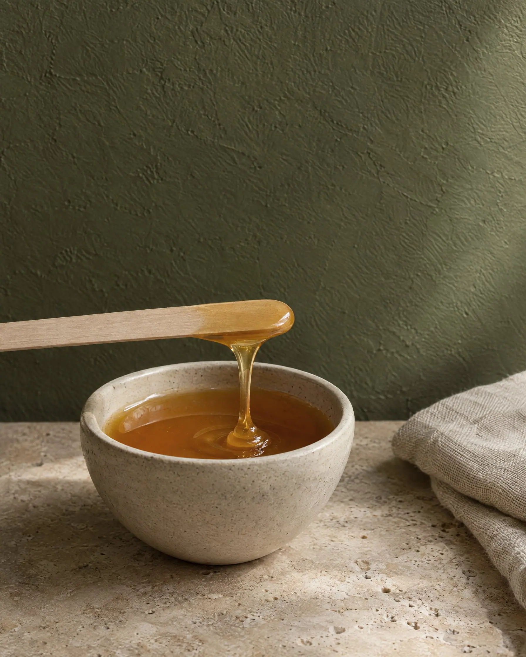 Conceptual image: a ribbon of warm amber wax falling from a wooden spatula into a stoneware bowl