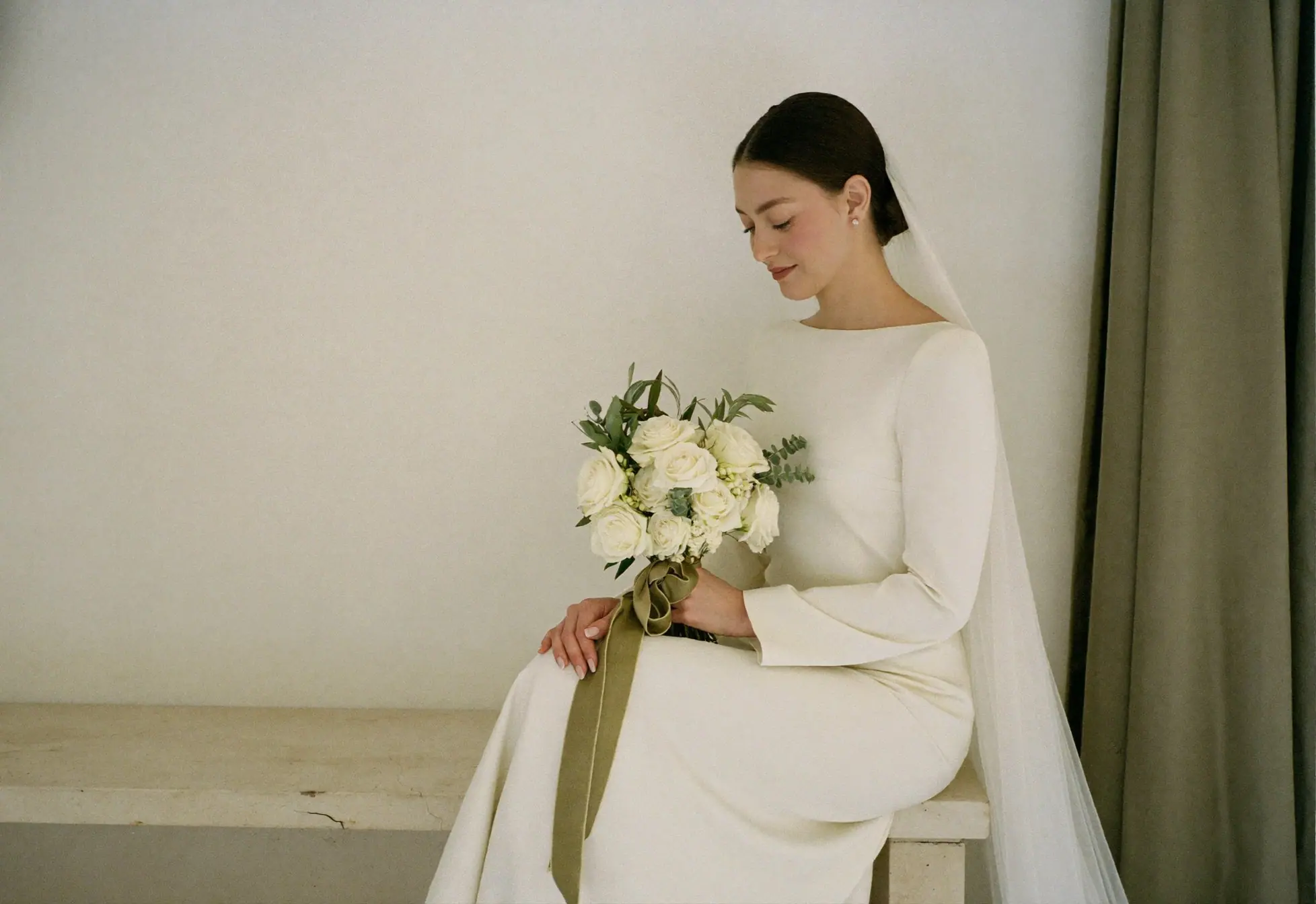 Conceptual image: a bride in an ivory gown and veil seated by a pale wall, holding ivory roses tied with an olive ribbon