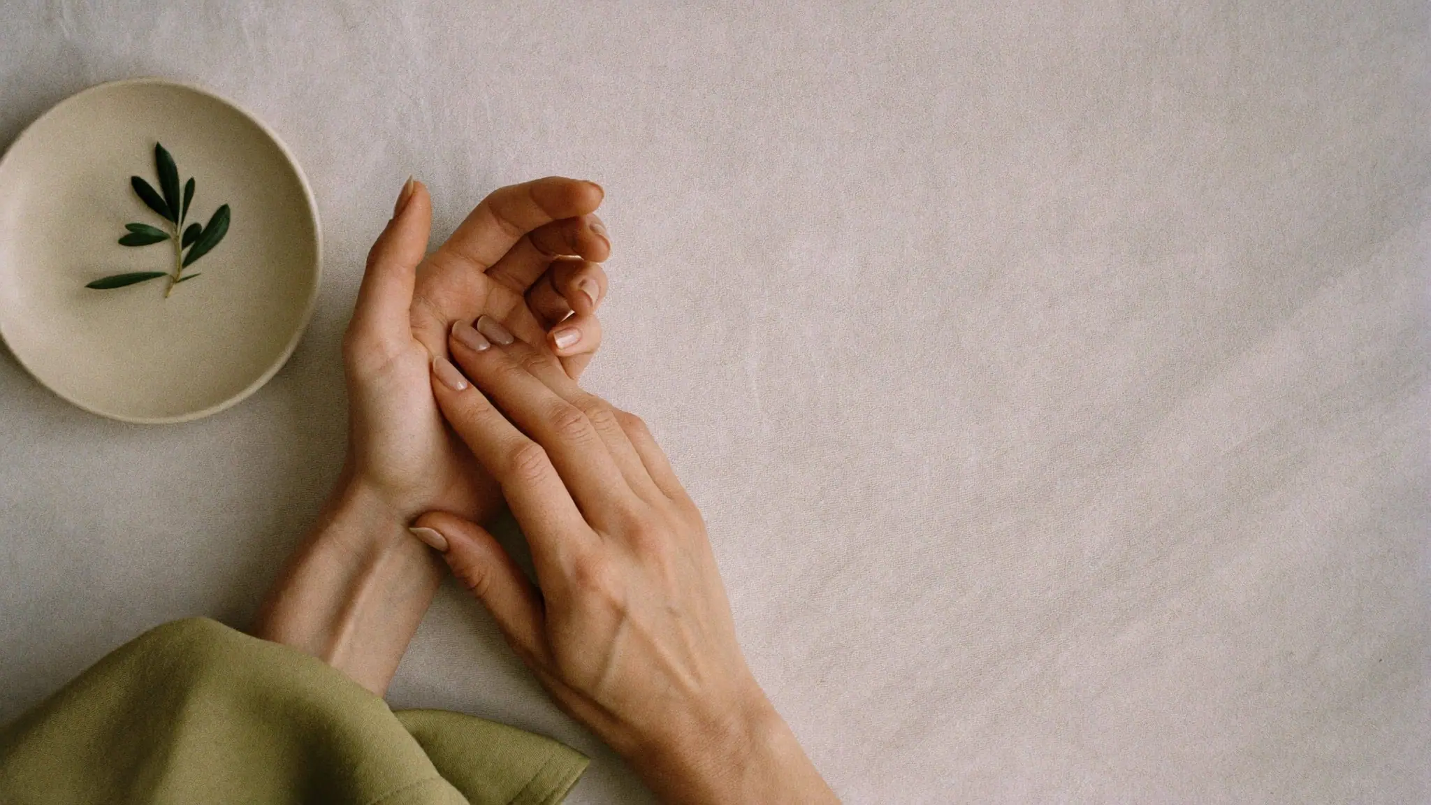 Conceptual image: overhead view of relaxed hands with sheer nude nails on ivory linen beside a small dish with an olive sprig