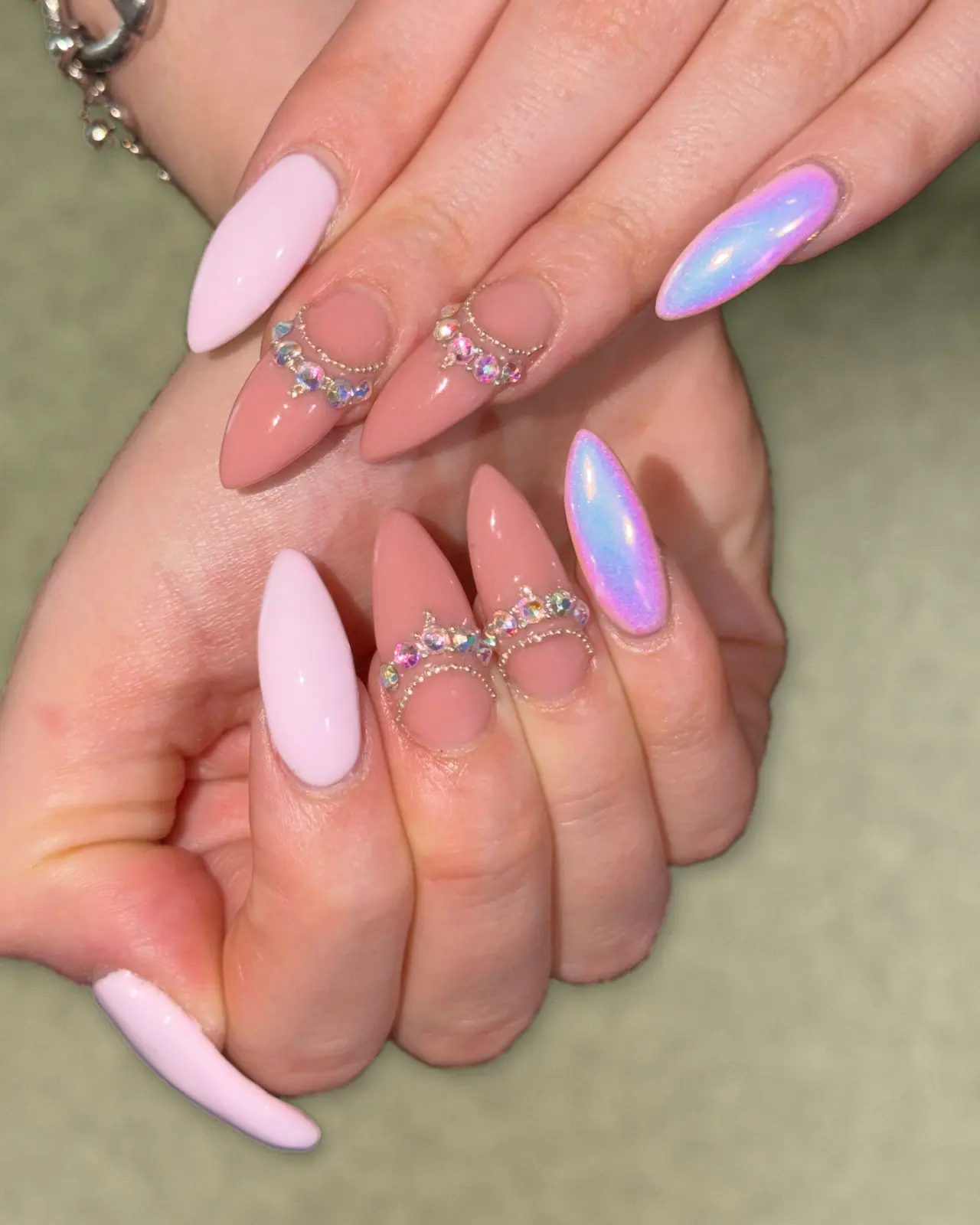 Two hands with long almond nails: soft pink, nude with rhinestone and bead crescents, and iridescent pink-blue chrome