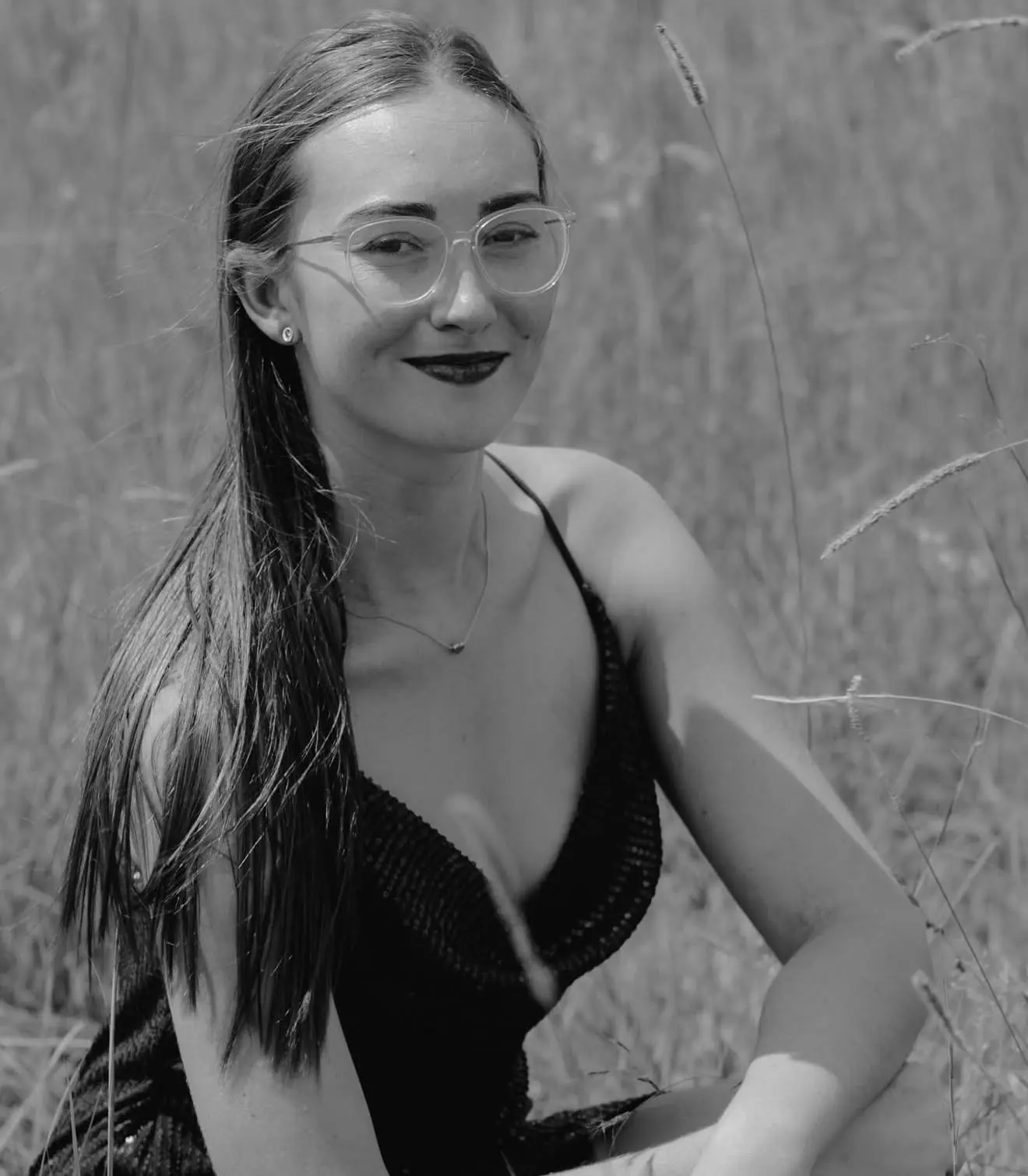 Black-and-white portrait of the owner of Rouxle Luxe Exclusive, smiling and wearing clear-framed glasses, seated in tall grass